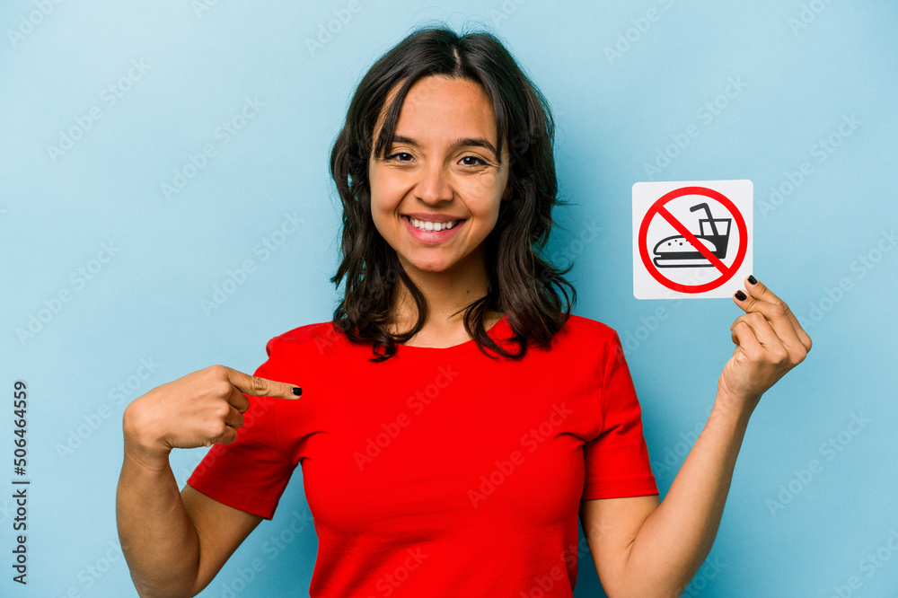 Young hispanic woman holding no eating sign isolated on blue background person pointing by hand to a shirt copy space, proud and confident Young hispanic woman holding no eating sign isolated on blue background person pointing by hand to a shirt copy space, proud and confident