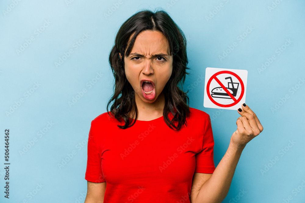 Young hispanic woman holding no eating sign isolated on blue background screaming very angry and aggressive. Young hispanic woman holding no eating sign isolated on blue background screaming very angry and aggressive.