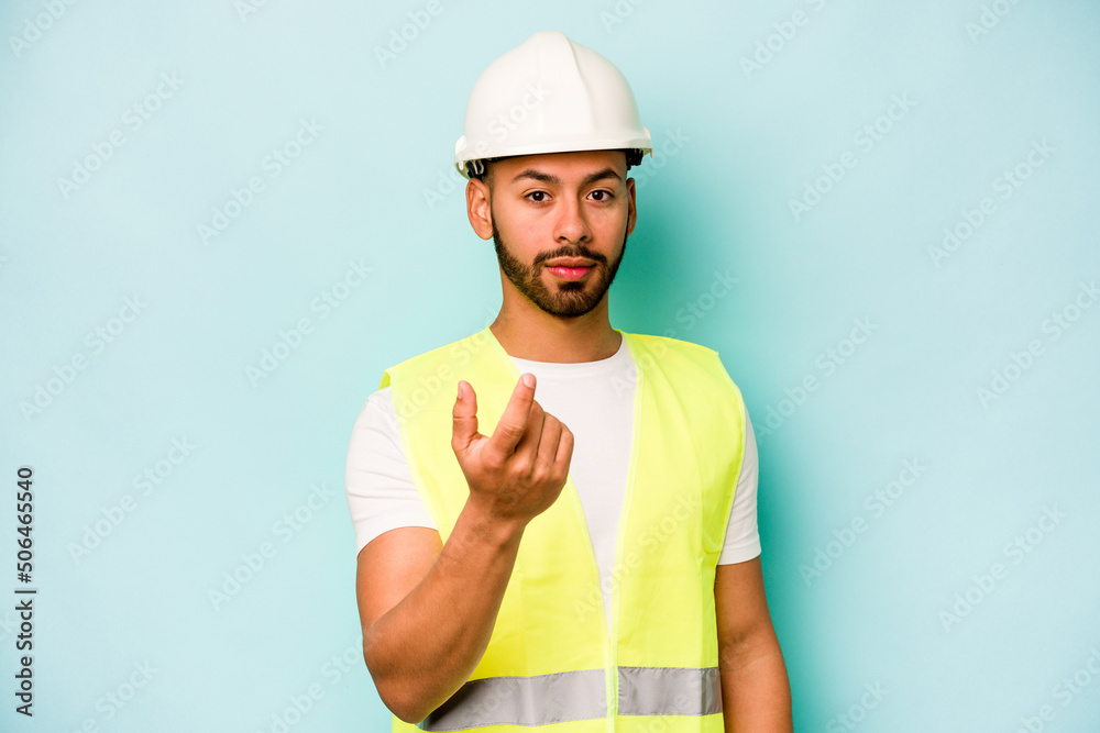 Young laborer hispanic man isolated on blue background pointing with finger at you as if inviting come closer. Young laborer hispanic man isolated on blue background pointing with finger at you as if inviting come closer.