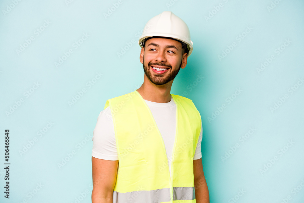 Young laborer hispanic man isolated on blue background looks aside smiling, cheerful and pleasant.