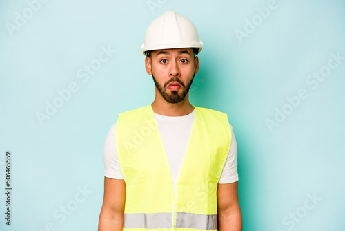 Canvas Print Young laborer hispanic man isolated on blue background shrugs shoulders and open eyes confused