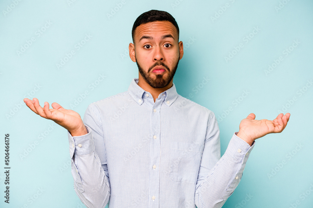 Young hispanic man isolated on blue background doubting and shrugging shoulders in questioning gesture.