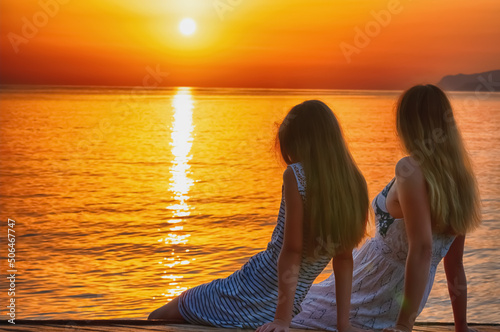 Rear view of mom and daughter sitting on a bridge by the sea and meeting morning sunrise. Summer holiday, rest, communication and travel concept. Horizontal image.