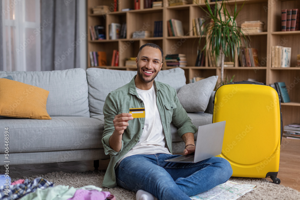 Cheerful black guy planning vacation trip, using computer and credit card to book hotel room online Cheerful black guy planning vacation trip, using computer and credit card to book hotel room online