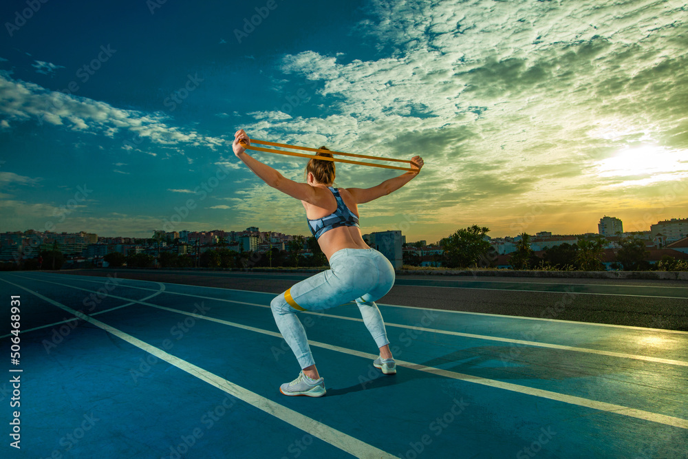 Workout with fitness bands. Sporty woman with slim tanned body in sportswear posing on sky background.