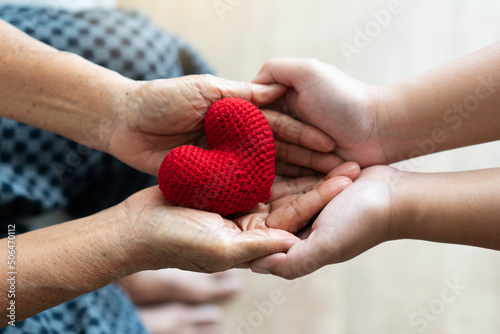 Young and senior woman holding each other hands and red yarn heart shape togetherness concept. Elderly care and protection with love from grandchild.