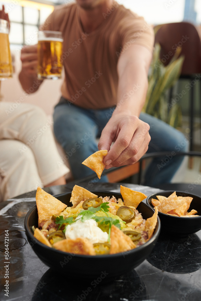 Man eating spicy nacho chips with guacamole and sour cream when drinking beer in bar with friends