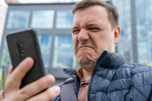 Caucasian man sad with what show on smartphone. Desperate sad annoyed millennial man looks at bad text message on mobile phone while standing outdoors