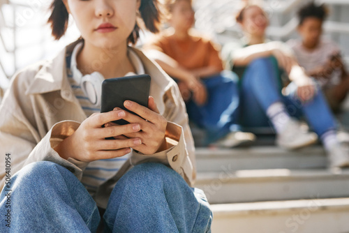 Closeup of teenage girl holding smartphone outdoors while sitting on metal stairs with group of friends in background, copy space