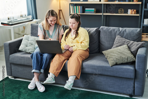 Young Caucasian woman wearing casual clothes sitting on couch teaching student with Down syndrome using laptop