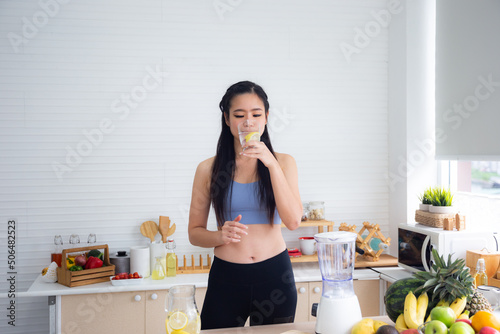 Fit and healthy young Asian woman in sportswear preparing and drinking fresh lemon water for healthy lifestyle and fitness in modern kitchen after morning workout and exercise