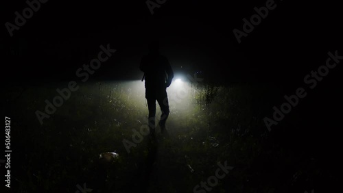 Dark male figure going through forest or countryside in light of car headlights at night. Search for missing person late at evening in natural area. Faceless man silhouette looking for someone photo