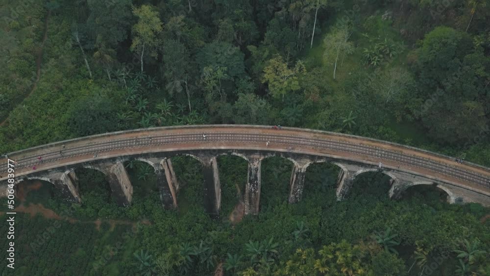 Drone aerial of famous Nine Arches Bridge Sri Lanka railway train with large old arches covered in moss tourist attraction deep in the tropical forest with groups of tourist walking along the track Drone aerial of famous Nine Arches Bridge Sri Lanka railway train with large old arches covered in moss tourist attraction deep in the tropical forest with groups of tourist walking along the track