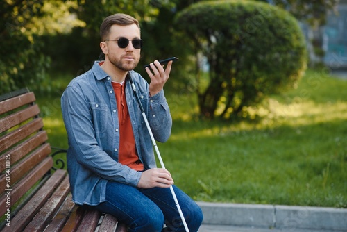 Fototapet Young blind man with smartphone sitting on bench in park in city, calling