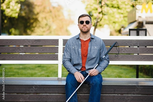 Visually impaired man with walking stick, sitting on bench in city park. Copy space photo