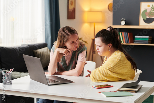 Young woman with Down syndrome and her university friend sitting at table working together on project looking at laptop screen and discussing issue at home photo