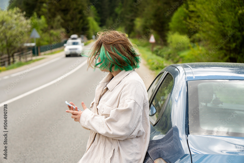 Caucasian hipster girl using a mobile phone to call the mechanic while looking at broken down car on street. Young girl with a broken car on the road call service. Cute girl near a broken car Caucasian hipster girl using a mobile phone to call the mechanic while looking at broken down car on street. Young girl with a broken car on the road call service. Cute girl near a broken car