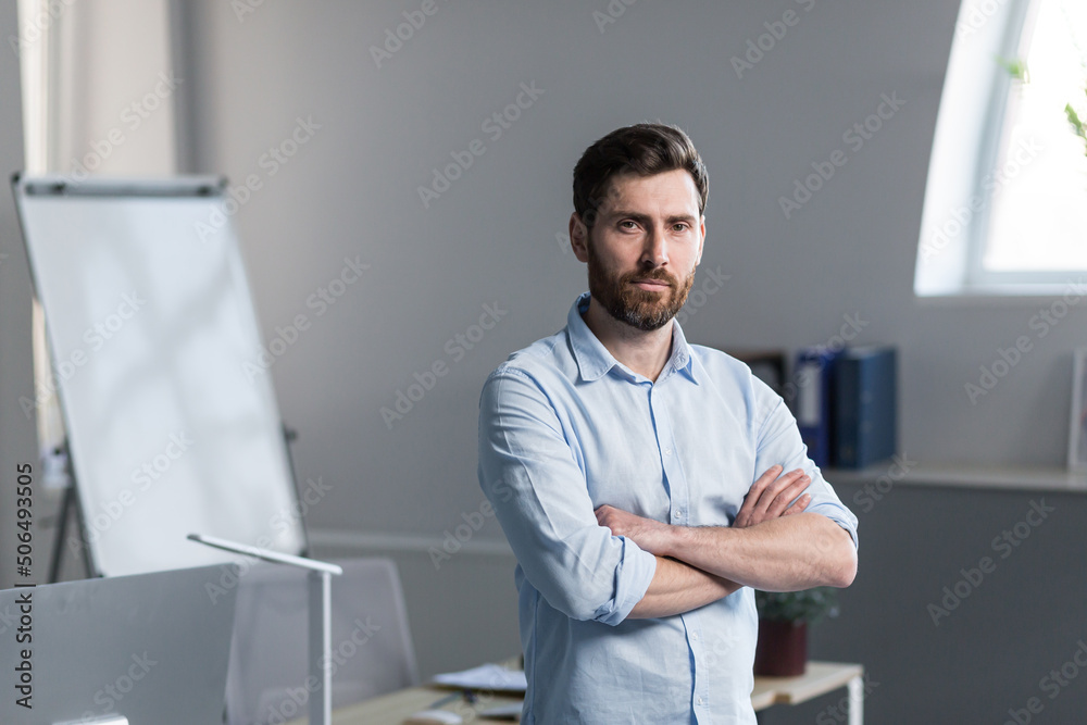 Close up photo. Portrait of a young man in the office, office worker. With a beard in a light shirt. He stands with his arms crossed. He looks at the camera, smiles