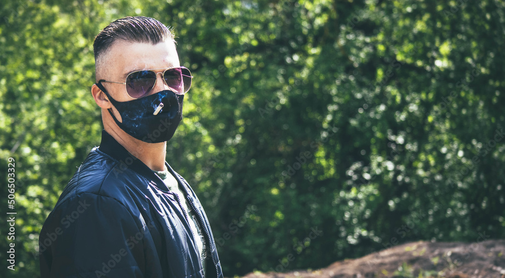 A man in a black jacket on the street smokes a cigarette through a hole in a protective mask. The guy in the black mask. High quality photo A man in a black jacket on the street smokes a cigarette through a hole in a protective mask. The guy in the black mask. High quality photo