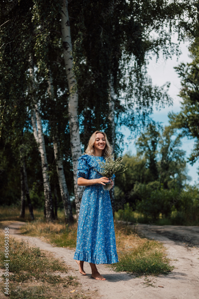 A young beautiful woman in a blue dress holds wild flowers in her hands