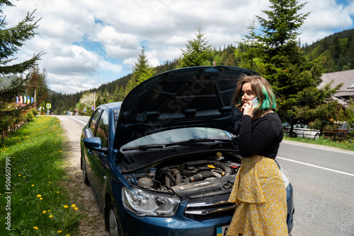 Caucasian hipster girl using a mobile phone to call the mechanic while looking at broken down car on street. Young girl with a broken car on the road call service. Cute girl near a broken car