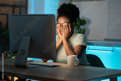 Symptoms of overworking. Young tired overworked african woman freelancer in eyeglasses working late from home, sitting in front of monitor, feeling eye strain and fatigue during computer work.  photo