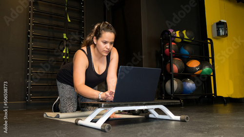 imágen de costado joven mujer en gimnasio hincada frente a   computadora prepara su clase online 