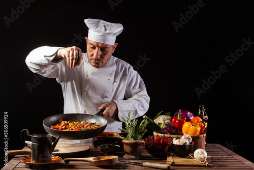 Male cook in white uniform and hat putting salt and herbs on food plate with vegetable