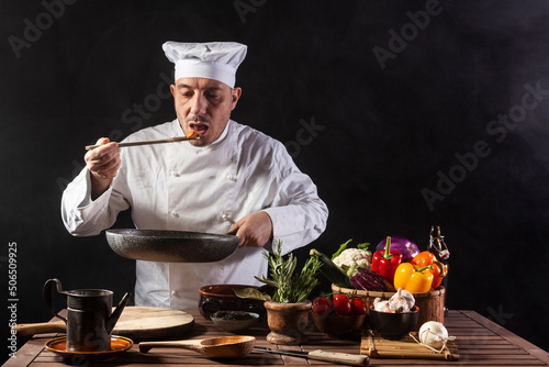 Male chef in white uniform is tasting food by using a wooden ladle at the kitchen of a restaurant photo