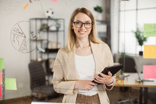 Portrait of beautiful young woman standing in front of glass board and smiling on camera. Female office worker in glasses and stylish suit using digital tablet in company. photo