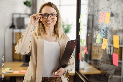Smiling business lady with clipboard standing near office glass wall with sticky notes and looking at camera. Female entrepreneur wearing stylish formal suit and eyeglasses. photo