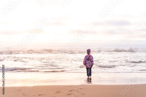 Lonely little girl standing on the ocean beach back to camera, looking on horizon. loneliness concept
