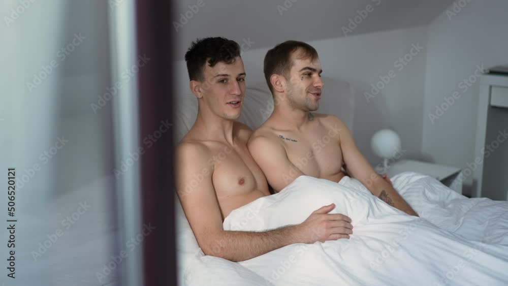 Shooting through window of happy relaxed gay couple sitting in bed talking in the morning. Confident relaxed Caucasian LGBTQ men chatting at home in bedroom indoors Shooting through window of happy relaxed gay couple sitting in bed talking in the morning. Confident relaxed Caucasian LGBTQ men chatting at home in bedroom indoors