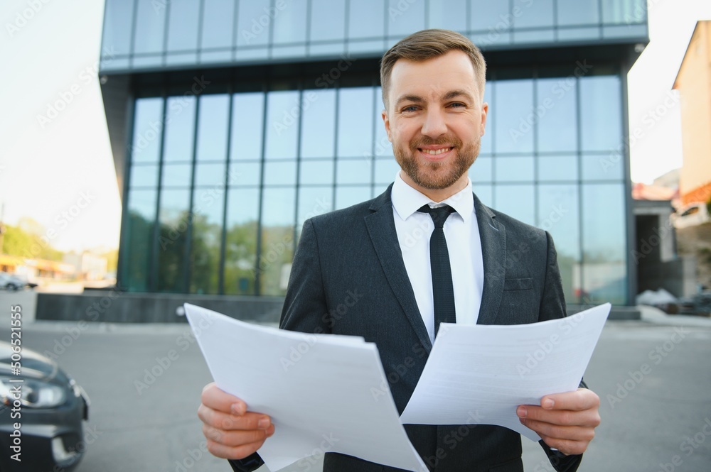 Portrait of handsome modern male businessman holding folder with documents on background of urban buildings and offices. Portrait of handsome modern male businessman holding folder with documents on background of urban buildings and offices.