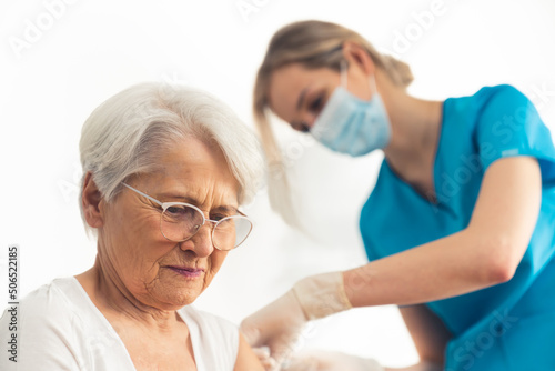 Elderly worried lady with grey hair sitting and having an injection in her arm done by a female nurse in a blue medical uniform. High quality photo