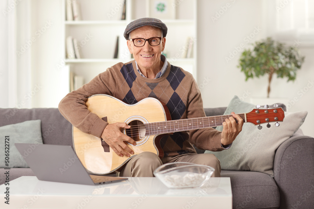 Pensioner playing an acoustic guitar and sitting on a sofa in front of a laptop computer