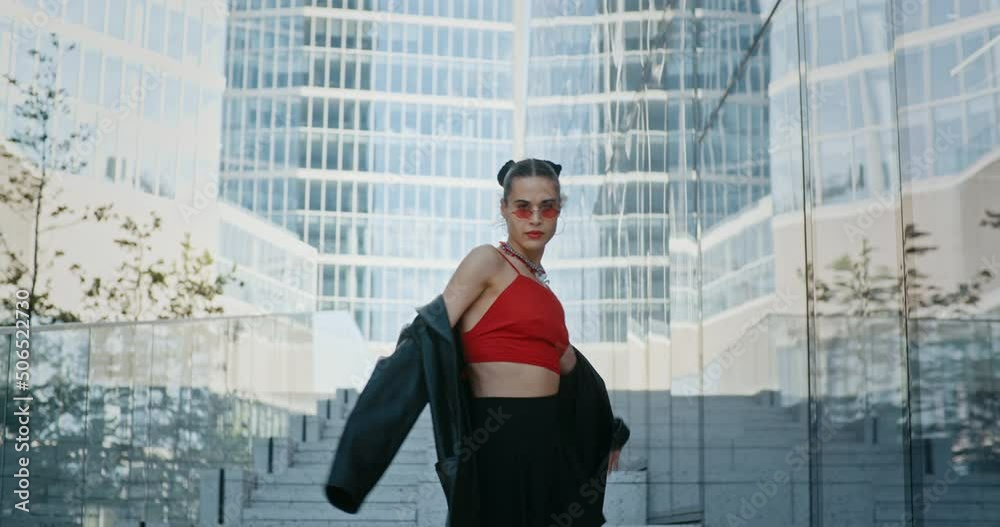 Gorgeous young woman, stylish cool fashion female model wearing leather jacket and black short skirt posing while standing on staircase in modern downtown high-rise glass skyscrapers in big city