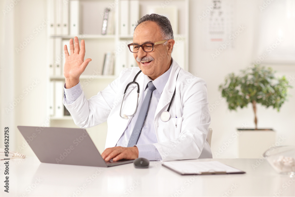 Mature male doctor sitting in an office and waving at a laptop computer