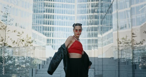 Gorgeous young woman, stylish cool fashion female model wearing leather jacket and black short skirt posing while standing on staircase in modern downtown high-rise glass skyscrapers in big city photo