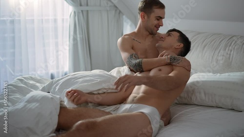 Focused young man surfing social media on smartphone as gay partner distracting hugging lying down on bed. Happy Caucasian LGBTQ couple cuddling in bedroom in the morning