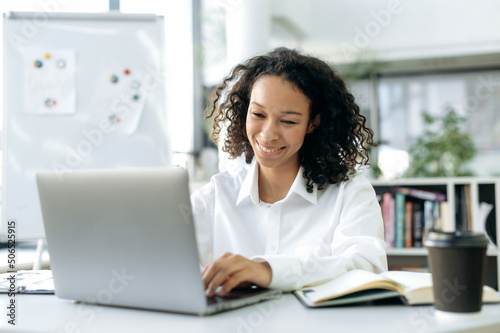 Charming African American girl, with curly hair, financial consultant, real estate agent, auditor, sits at desk in front of a laptop, chats with colleagues or employees, looks at the screen, smiles