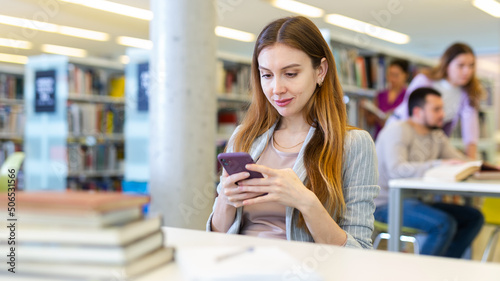 Positive relaxed young female student sitting with smartphone in hands at table in university library  taking break from study..