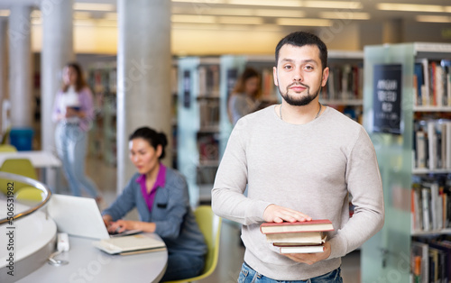 Portrait of a confident student guy, standing in the university library, holding textbooks in his hands