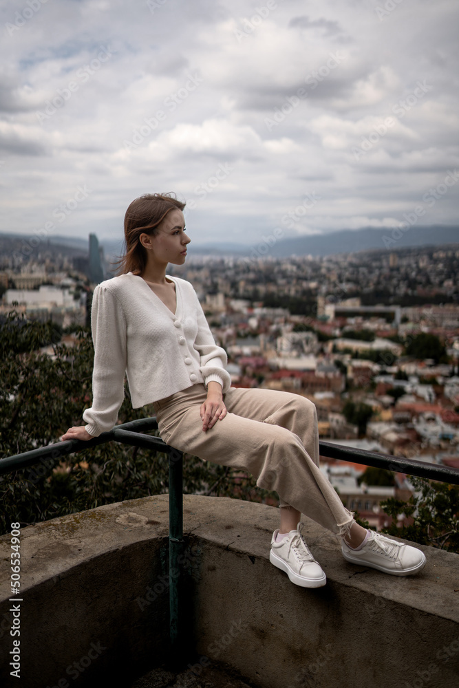 Stylish woman in a white sweater and beige pants is posing on the background of the city.