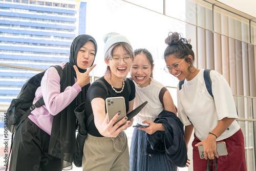Fototapeta Group of young happy Asian women with diverse ethnicity laughing having fun while taking selfie with a smartphone