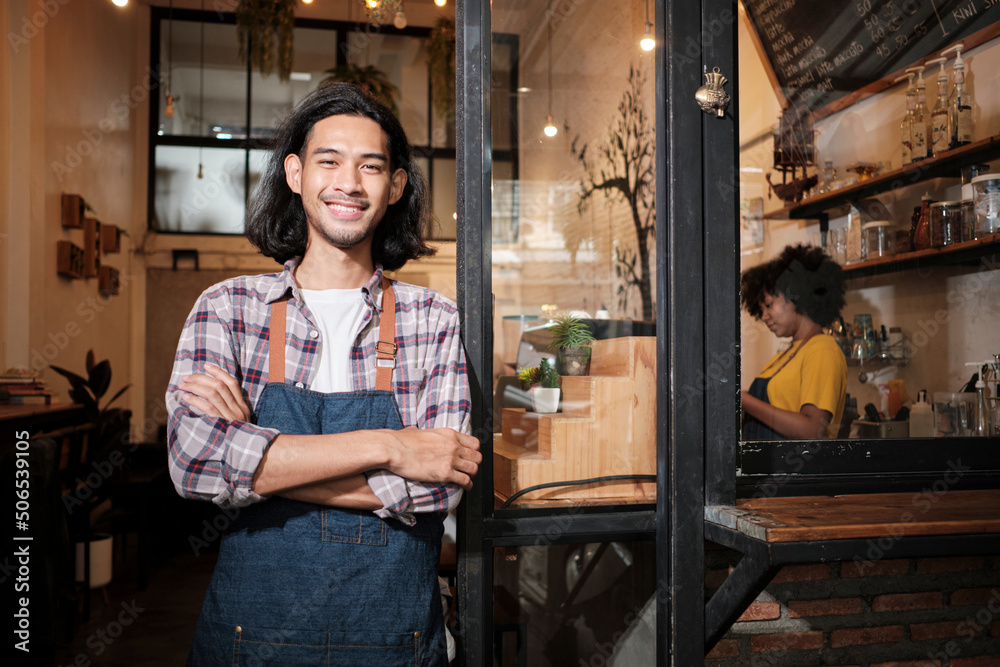 A long hair handsome Asian male startup barista with apron standing at casual cafe door, arms crossed, looking at the camera with and welcoming smile, happy and cheerful with coffee shop service jobs.