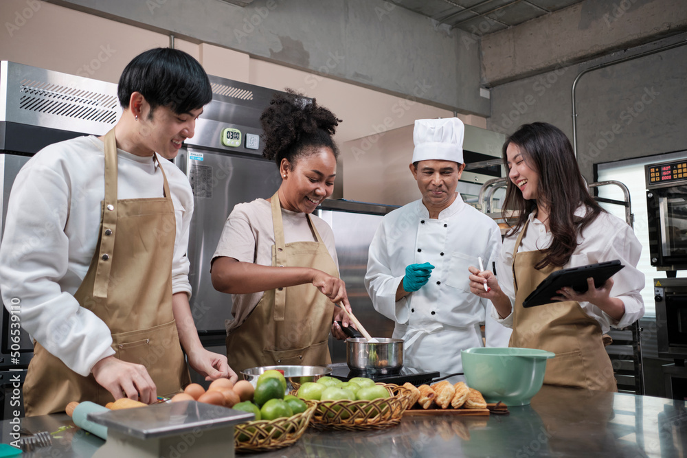 Hobby cuisine course, senior male chef in cook uniform teaches young cooking class students to prepare, mix and stir ingredients for pastry foods, fruit pies in restaurant stainless steel kitchen. Hobby cuisine course, senior male chef in cook uniform teaches young cooking class students to prepare, mix and stir ingredients for pastry foods, fruit pies in restaurant stainless steel kitchen.