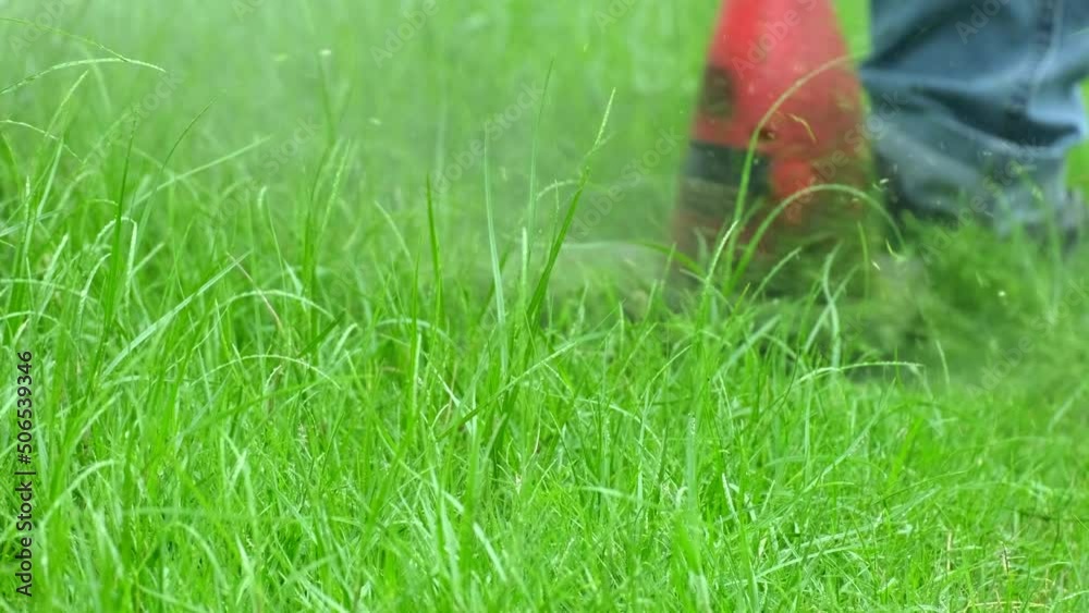 Close-up shot of male worker in jeans maintenance garden using an electric lawnmower to trim overgrown green grass in the lawn yard, scattering the grass in summer, outdoor landscaping work service. Close-up shot of male worker in jeans maintenance garden using an electric lawnmower to trim overgrown green grass in the lawn yard, scattering the grass in summer, outdoor landscaping work service.