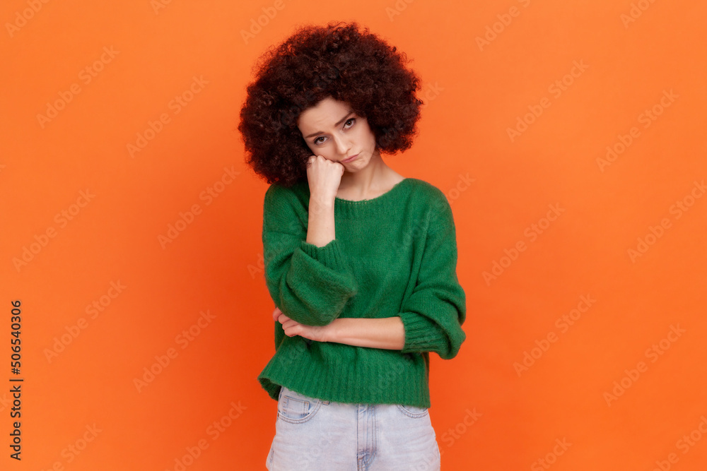 Portrait of bored woman with Afro hairstyle wearing green casual style sweater standing with fist under chin, listening to boring story. Indoor studio shot isolated on orange background. Portrait of bored woman with Afro hairstyle wearing green casual style sweater standing with fist under chin, listening to boring story. Indoor studio shot isolated on orange background.