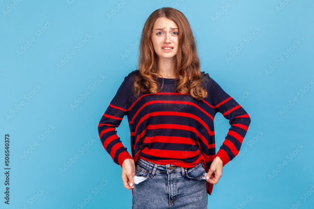 I'm bankrupt! Unhappy poor woman, turning empty pockets inside out and looking frustrated by overspend, lack of money, wearing striped casual sweater. Indoor studio shot isolated on blue background. I'm bankrupt! Unhappy poor woman, turning empty pockets inside out and looking frustrated by overspend, lack of money, wearing striped casual sweater. Indoor studio shot isolated on blue background.